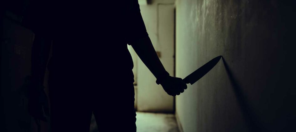 Person geht mit Messer in der Hand durch dunklen Flur.
